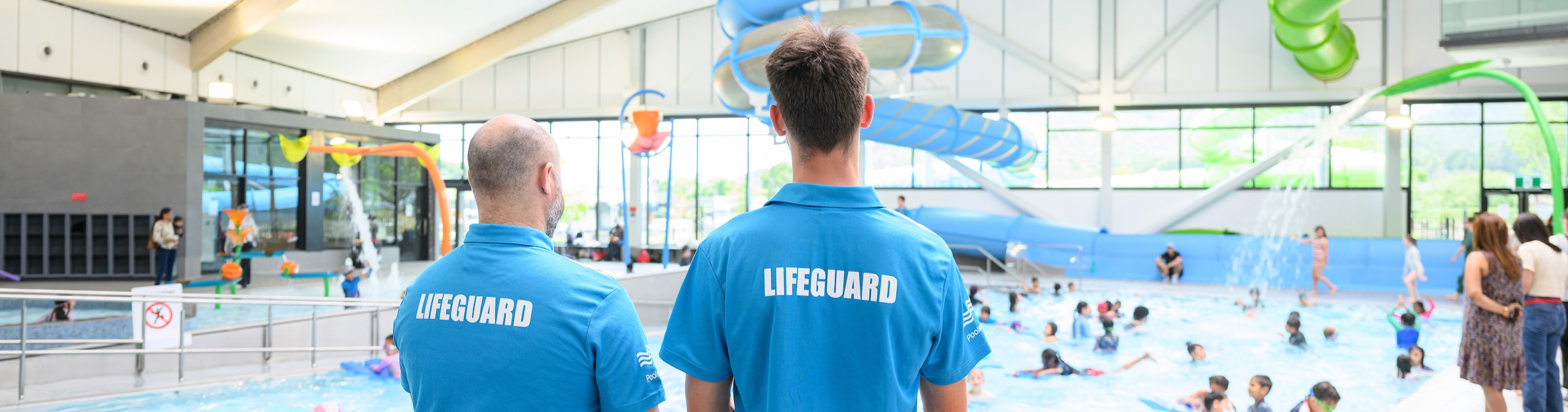 Two lifeguards stand with their backs to the camera watching over children playing in a pool